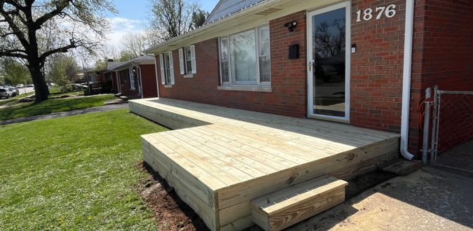 A newly built wooden deck with light-colored planks and low steps, extending from the side of a red brick house with a glass door, set in a sunny backyard with green grass.